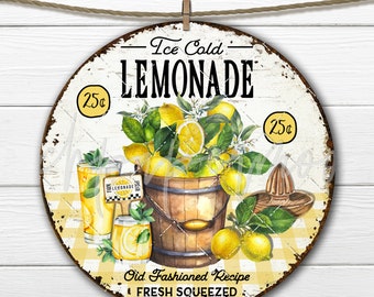 May include: A round, white metal sign with a yellow and black checkered background. The sign says "Ice Cold Lemonade" in black text with a yellow circle around the text "25c". There are lemons, a bucket of lemons, and two glasses of lemonade on the sign.