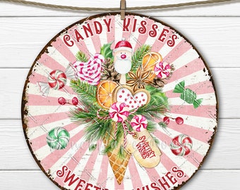 May include: A pink and white round sign with a red and white striped background. The sign says "Candy Kisses Sweetest Wishes" in red letters. The center of the sign features a watercolor illustration of a cone filled with candy canes, pinecones, oranges, and a Santa Claus figurine.
