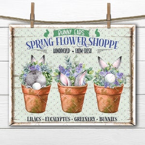 Peut inclure: Une enseigne de style vintage avec le texte "Bunny Ears Spring Flower Shoppe" et les mots "Handpicked - Farm Fresh" en dessous. L'enseigne présente trois plantes en pot avec des oreilles de lapin qui dépassent du haut. Les plantes sont étiquetées "Lilacs - Eucalyptus - Greenery - Bunnies".