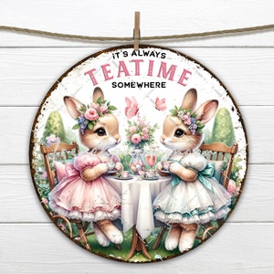 Op de afbeelding: Een rond metalen bord met een witte achtergrond en een verweerde look. Het toont twee konijnen die bloemenkransen en jurken dragen, zittend aan een tafel met theekopjes en een theepot. Het bord zegt "It's Always Teatime Somewhere".