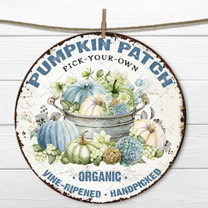 May include: A round metal sign with a white background and blue text that reads "Pumpkin Patch Pick-Your-Own Organic Vine-Ripened Handpicked". The sign features a watercolor illustration of a galvanized bucket overflowing with blue, green, and white pumpkins surrounded by white and blue flowers.