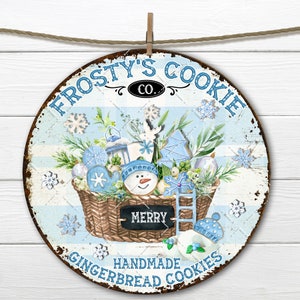May include: Circular metal sign with a distressed finish, depicting a winter scene with gingerbread cookies in a basket. The sign reads "Frosty's Cookie Co." and "Handmade Gingerbread Cookies". The design includes snowflakes and a snowman cookie.