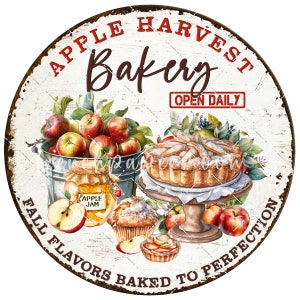 May include: A round, white sign with red and brown text that reads "Apple Harvest Bakery Open Daily Fall Flavors Baked to Perfection". The sign features a watercolor illustration of apples, a pie, and other baked goods.