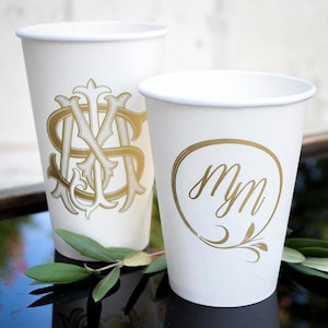May include: Two white paper cups featuring gold foil designs. The taller cup displays an ornate, intertwined monogram of the letters M and S. The shorter cup features a circular gold frame enclosing the cursive, lowercase letters mm with a leaf accent.