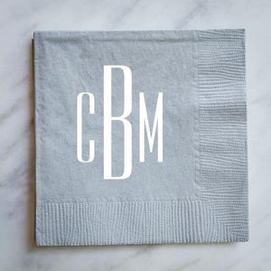 May include: A square light gray paper napkin featuring a textured border and a large white monogram in the center displaying the letters CBM in a tall, clean, sans-serif font. The napkin has a matte finish and a distinct embossed edge pattern.