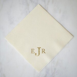 May include: A square, cream-colored paper napkin featuring a centered, gold-colored monogram with the letters E, J, and R. The letter J is larger than the letters E and R, which are positioned on either side of the central character.