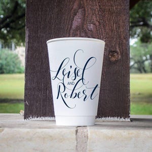 May include: A white disposable paper cup featuring dark blue cursive calligraphy text that reads Leisel AND Robert. The cup has a smooth, tapered cylindrical shape and is positioned upright, displaying the printed names clearly on the front surface.