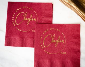 May include: Two burgundy paper napkins with gold lettering. The napkins feature the names "Brittni and Dylan" and "Clayton" in a circular design, with the date "11.9.19" below. The napkins are folded and have a textured surface.