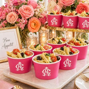 May include: Several bright pink paper cups filled with pasta salad containing penne, cherry tomatoes, and black olives. Each cup features a white monogram logo with the letters AG. The cups are arranged on a white marble serving tray.