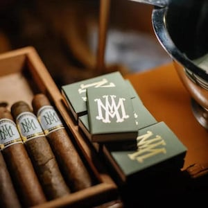 May include: A wooden cigar box with several cigars and a stack of green matchboxes. The matchboxes have a gold monogram on them. The cigars have a white band with a monogram. The scene is set on a wooden surface.