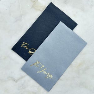 May include: Two rectangular paper napkins, one dark navy blue and one light grey, both featuring gold foil script lettering. The navy napkin displays the text The Q, while the light grey napkin features the text The Youngs in elegant cursive.