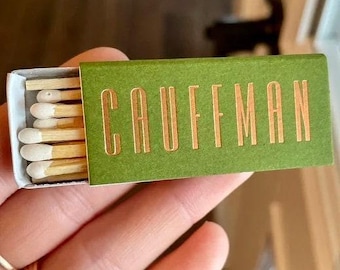 May include: A rectangular matchbox with a matte olive green cover featuring the word CAUFFMAN printed in metallic gold capital letters. The box is partially open, revealing several wooden matches with white tips inside the white cardboard tray.