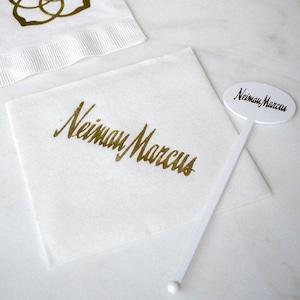May include: A white square paper napkin featuring the gold foil text Neiman Marcus printed diagonally across the center, accompanied by a white plastic stirrer with an oval top displaying the black text Neiman Marcus and a partial napkin.