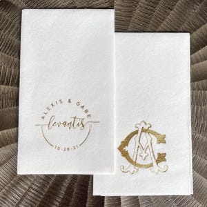 May include: Two rectangular white paper napkins featuring gold foil stamping. One napkin displays the text Alexis & Gabe, the surname Levantis, and the date 10.28.21 within a circular border. The other napkin features a large gold monogram of the letters A and C.