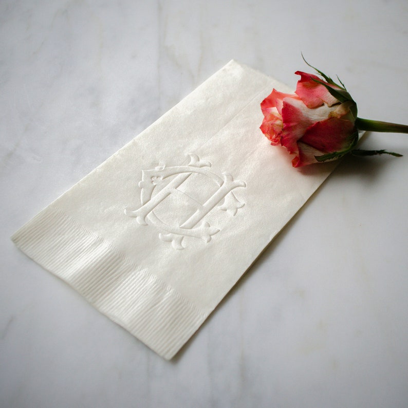 May include: A white linen napkin with an embossed monogram of the letter "H" in a decorative frame. A single pink rose is placed on the napkin.
