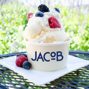 May include: A scoop of vanilla ice cream in a cream-colored paper cup with the text JACOB printed in dark blue. The ice cream is topped with whipped cream, fresh blackberries, blueberries, and raspberries, with additional berries on the plate.