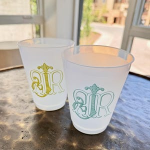 May include: Two translucent frosted plastic cups featuring decorative monograms. The left cup displays a gold-colored letter design, while the right cup features a teal-colored letter design. Both cups have a simple, tapered cylindrical shape.