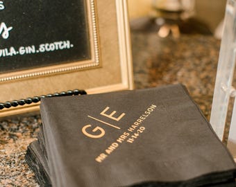 May include: A stack of square black paper napkins featuring gold foil lettering. The design includes the initials G/E, the text MR AND MRS HARRELSON, and the date 11-14-20 printed in a clean, sans-serif font on the top napkin surface.
