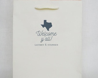 May include: A cream-colored paper gift bag with white twisted rope handles and pale pink tissue paper peeking from the top. The front features a dark blue silhouette of the state of Texas above the text Welcome y'all! and LAUREN & STEPHEN.