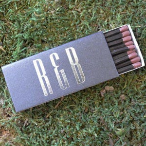 May include: A rectangular matchbox with a dark gray exterior featuring the silver text R&B printed on the front. The box is partially open, revealing a row of matches with dark brown wooden sticks and light pink match heads inside.