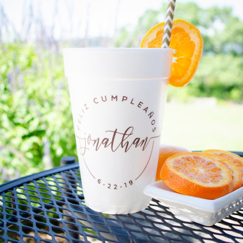 May include: White foam cup with a brown circle design that says "Feliz Cumpleanos Jonathan 6.22.19". The cup is sitting on a black metal table with orange slices on a white plate.