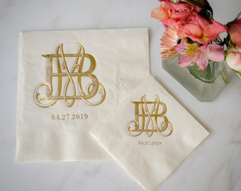 May include: Two cream-colored paper napkins featuring a gold foil-stamped monogram of the letters B and M intertwined with elegant flourishes. Below the monogram, the date 04.27.2019 is printed in gold. The napkins have a textured border edge.