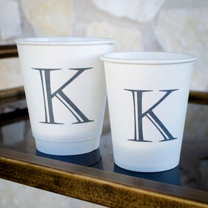 May include: Two white paper cups with a gray letter K printed on each. The cups are on a gold metal surface.