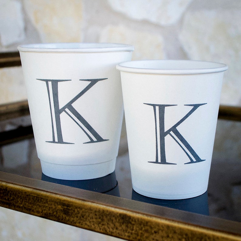 May include: Two white paper cups with a gray letter K printed on each. The cups are on a gold metal surface.