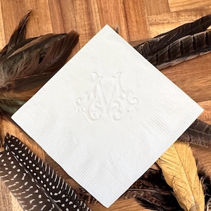 May include: A square white paper napkin featuring an embossed letter M in the center with decorative scrollwork details. The napkin has a textured border and is positioned diagonally, showcasing a crisp, clean finish and a formal design.