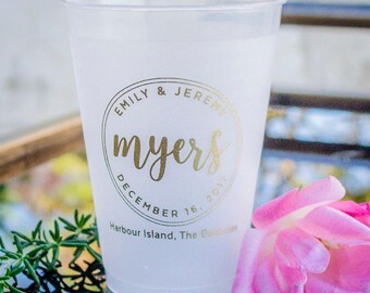 May include: A frosted plastic cup featuring gold foil text that reads Emily & Jeremy, myers, December 16, 2017, and Harbour Island, The Bahamas. The cup is decorated with a circular emblem design and is positioned next to a pink flower.
