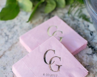 May include: Pink paper napkins with gold-colored initials "CG" and the date "6-10-2017" printed on them. The napkins are stacked on a light-colored surface, with white roses and greenery in the background.
