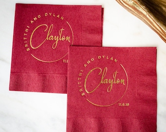 May include: Two square deep red paper napkins featuring gold foil stamping. The design includes a circular border with the text "BRITTNI AND DYLAN" arched above, the name "Clayton" in a script font in the center, and the date "11.9.19" below.