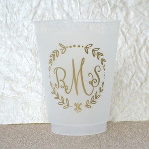May include: A white plastic cup with a gold monogram design. The monogram is "BMS" surrounded by a wreath of leaves.