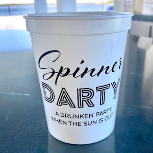 May include: A white plastic cup featuring black text that reads Spinner DARTY A DRUNKEN PARTY WHEN THE SUN IS OUT. The word Spinner is in a cursive script font, while DARTY is in a bold, outlined block font, and the remaining text is in sans-serif.
