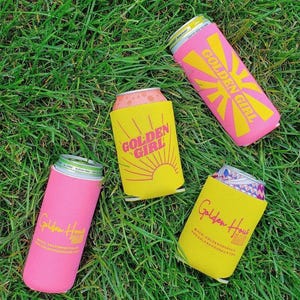 May include: Four beverage can coolers in pink and yellow, with the text "Golden Girl" and "Golden Hour" printed on them. The coolers are on a bed of green grass.