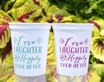 May include: Two white plastic cups featuring the text Love Laughter and Happily Ever After. One cup has the text printed in teal, while the other cup features the same text printed in a vibrant purple color. Both cups have a smooth finish.