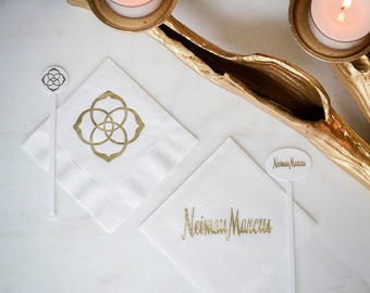 May include: Two white paper napkins and two white plastic stirrers. One napkin features a gold Kendra Scott logo, the other features gold Neiman Marcus text. The stirrers match the respective logos. A gold candle holder with two lit tea lights is visible.