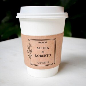 May include: A white disposable coffee cup with a white plastic lid and a tan cardboard sleeve. The sleeve features a black rectangular border, a botanical line drawing, and the text RAMOS, ALICIA & ROBERTO, and the date 12-06-2025 printed in black.