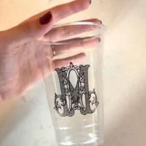 May include: A clear plastic disposable cup featuring a black decorative monogram of the letter M. The cup has a smooth, cylindrical body with a slightly wider rim at the top and a tapered base, showcasing a detailed, ornate vintage-style font.