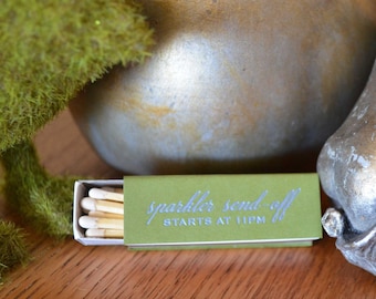 May include: A rectangular olive green matchbox containing wooden matches with white tips. The front of the box features the silver text "sparkler send-off STARTS AT 11PM" printed in a combination of script and sans-serif fonts.