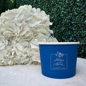 May include: A deep blue paper cup featuring a white square frame with a floral illustration at the top, the names Patricia and Marcus in script, and the date December 11, 2026, printed below. A large cluster of white hydrangea flowers sits behind.