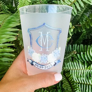 May include: A frosted plastic cup featuring a blue crest with the monogram letters MC, decorated with light blue and white floral accents and a blue ribbon banner at the bottom containing the printed date 09.09.2023 in black text.