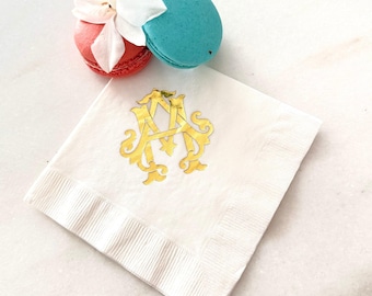 May include: A square white paper napkin featuring a gold foil-stamped monogram of the letters A and M. Positioned above the napkin are a red macaron and a blue macaron, with a small white flower petal resting between the two colorful cookies.