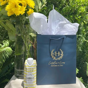 May include: A dark blue gift bag with gold lettering reading "CHL", "Caroline & Lucas", and "OCTOBER 16, 2021" with white tissue paper, next to a carton of Coco Luxe Boost pure coconut water and a clear glass vase filled with yellow flowers.