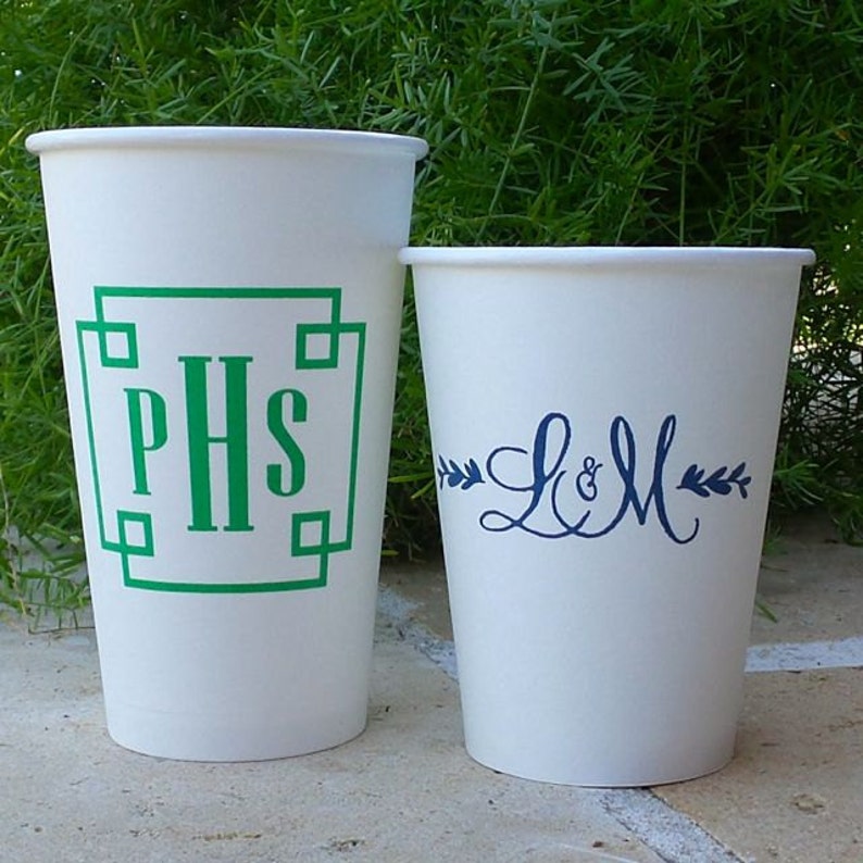 May include: Two white paper cups with personalized designs. The first cup has a green monogram "PHS" in a square frame. The second cup has a blue cursive monogram "L&M" with green leafy branches on either side.