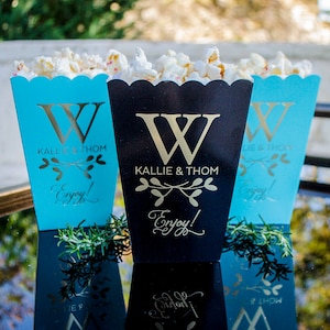 K&ouml;nnte beinhalten: Drei Popcorn-Boxen, eine schwarze und zwei t&uuml;rkisfarbene, gef&uuml;llt mit Popcorn. Jede Box hat einen goldenen "W"-Initialen, die Namen "Kallie & Thom" und das Wort "Enjoy!" in goldener Schrift. Die Boxen sind auf einer reflektierenden Oberfl&auml;che angeordnet.