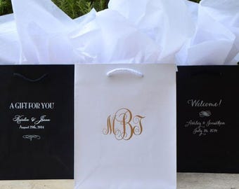 Pode incluir: Três sacos de presente com papel de seda branco. Dois sacos pretos apresentam texto branco com A GIFT FOR YOU, Kristin & Jason, August 29th, 2014 e Welcome!, Ashley & Jonathon, July 26, 2014. Um saco branco central exibe um monograma dourado com as letras MBJ.