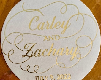 May include: A circular white coaster featuring gold foil lettering. The text reads Carley AND Zachary in a decorative script font, with the date JULY 9, 2022 printed in a smaller, simple serif font below the names, surrounded by gold flourishes.