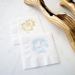 May include: Two square white paper napkins with embossed borders. One napkin features a gold foil monogram with the letters VS in an ornate script. The second napkin displays a light blue monogram with the letter C in a decorative scroll design.