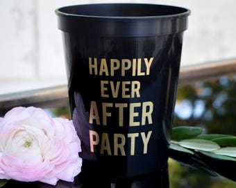 May include: A black plastic stadium cup featuring the gold text "HAPPILY EVER AFTER PARTY" printed on the front. The cup is positioned next to a light pink flower with layered petals and green leaves, reflecting on a dark surface.